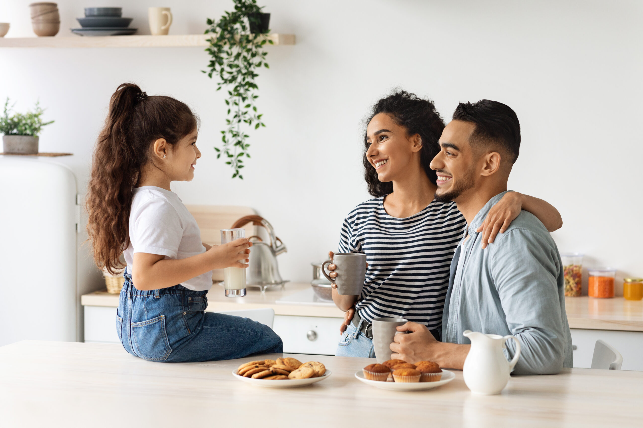 Happy arab family enjoying breakfast at cozy kitchen, little girl daughter sitting on table, drinking milk and having conversation with hugging smiling father and mother drinking morning coffee